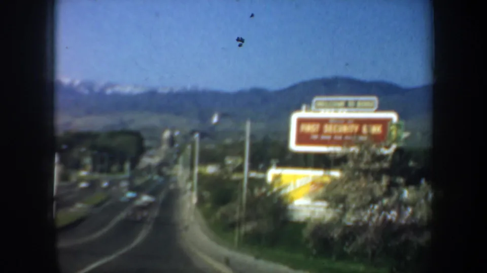 Cataloged Oregon 1957 road footage with cars on a broad street, a red First Security Bank sign, and mountains rising in the background.