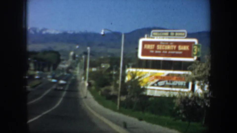 A closer 1957 drive-by frame shows the First Security Bank sign, roadside planting, light traffic, and a mountain ridge beyond town.