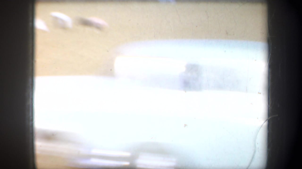A soft, overexposed Oregon 1957 home-movie frame showing a pale passing vehicle or object against dry open ground.
