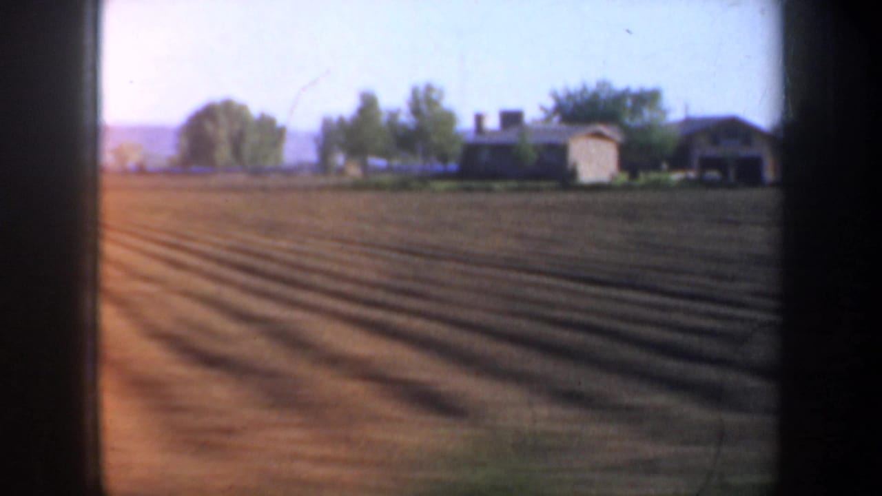 Passing 1957 Oregon farmland view with brown furrows, a small house or farm structure, trees, and low hills beyond.