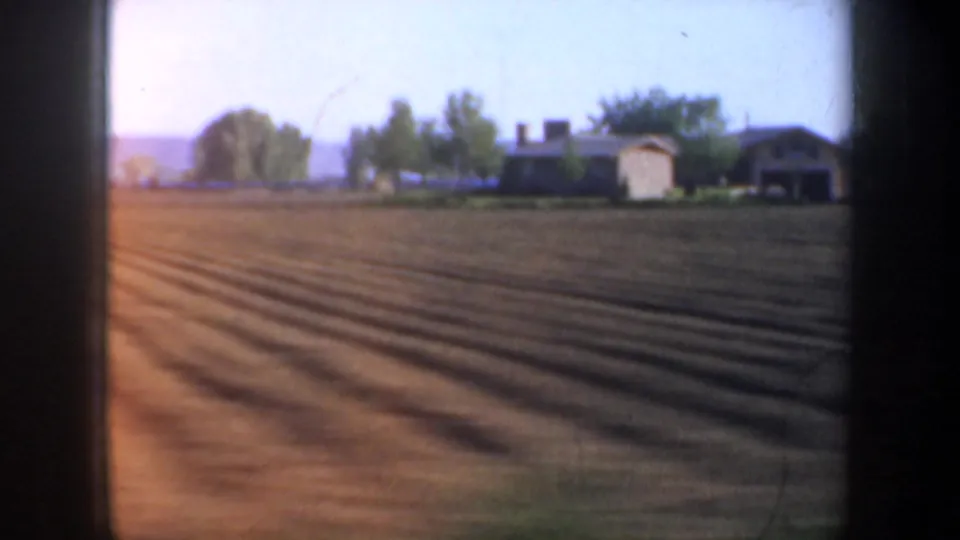 Passing 1957 Oregon farmland view with brown furrows, a small house or farm structure, trees, and low hills beyond.