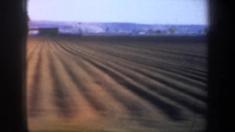 1957 Oregon 8mm roadside view of long brown field rows extending toward a pale horizon and distant hills.