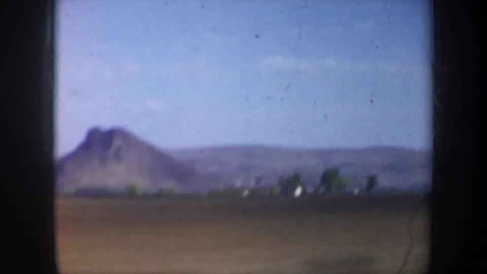 Final 1957 Oregon frame showing open farmland, a prominent distant hill or butte, low hills, and small pale structures or highlights near the horizon.