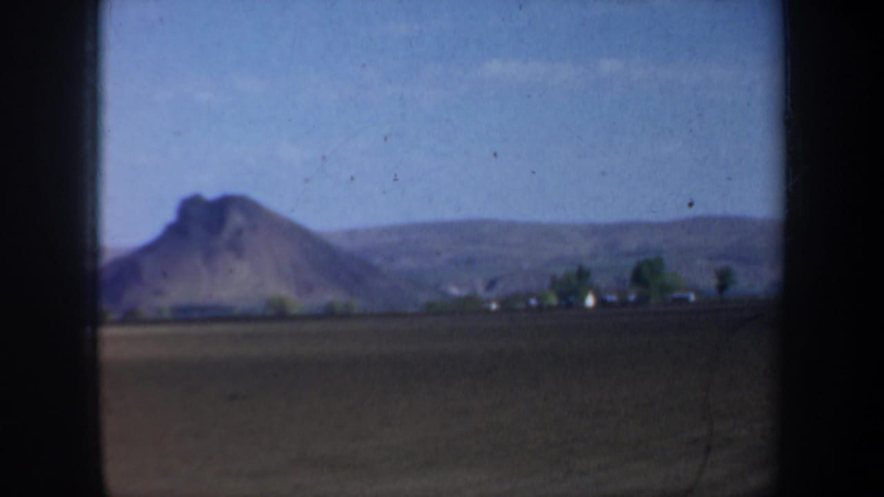 1957 Oregon 8mm frame showing a rural field, rocky hill or mountain, low houses, trees, and blue sky from a moving-car viewpoint.
