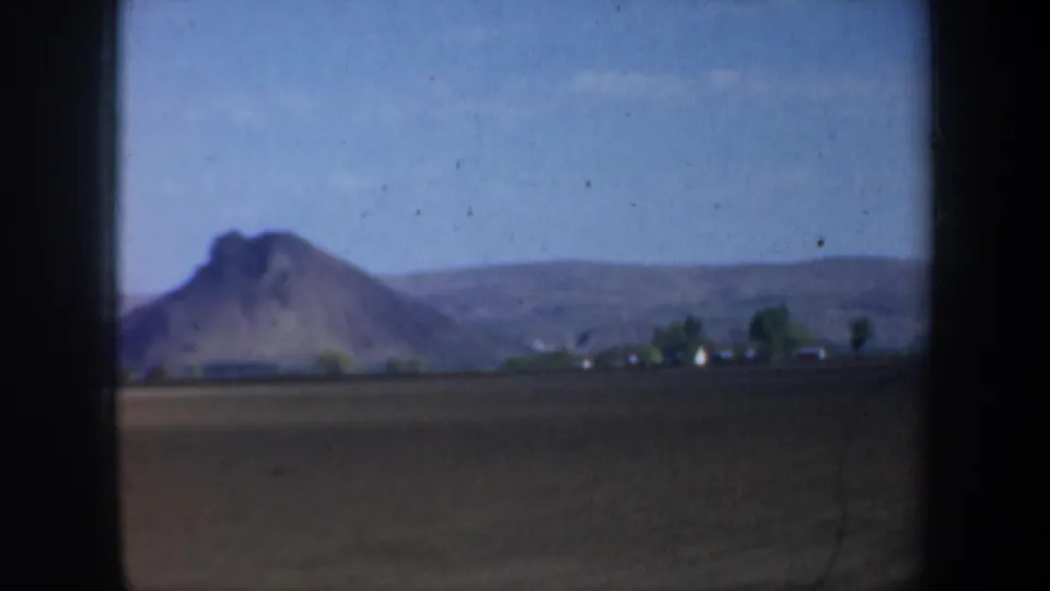 1957 Oregon 8mm frame showing a rural field, rocky hill or mountain, low houses, trees, and blue sky from a moving-car viewpoint.