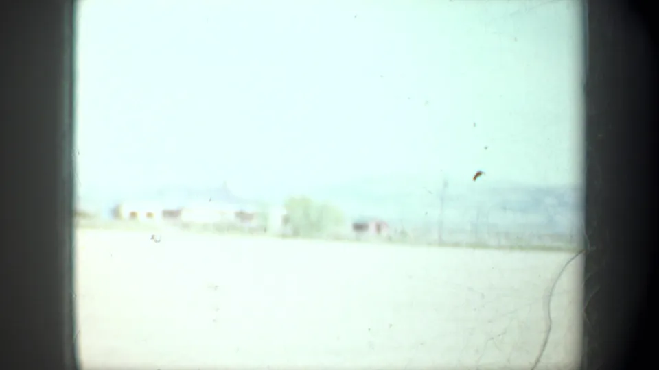 Bright 1957 Oregon roadside frame with washed-out field, faint distant buildings, trees, and a pale sky seen from a moving vehicle.