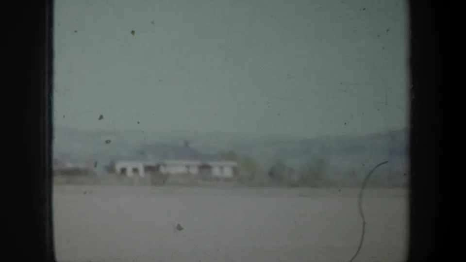 Soft 8mm Oregon landscape frame with a pale field, low rural buildings, distant hills, scattered trees, and visible film wear.