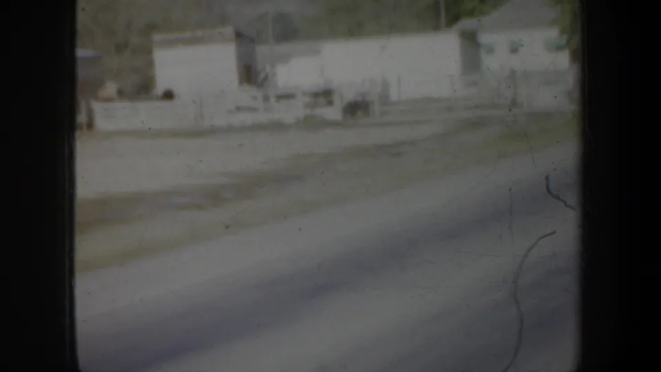 1957 Oregon 8mm frame showing low roadside buildings, pavement, scrubby vegetation, and dark film edges from a moving-car perspective.