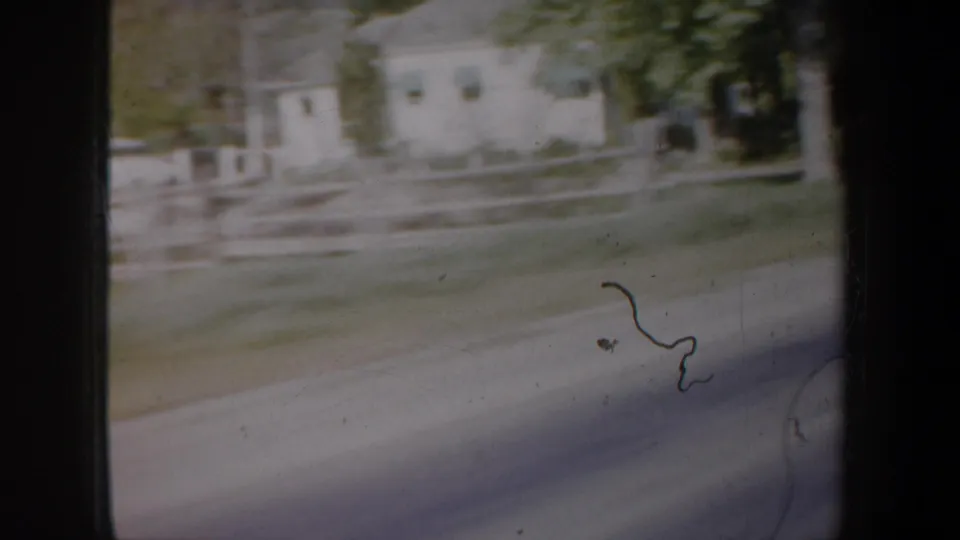 1957 Oregon road-view frame with a low light-colored building, fencing, trees, and road blur through the vehicle window.