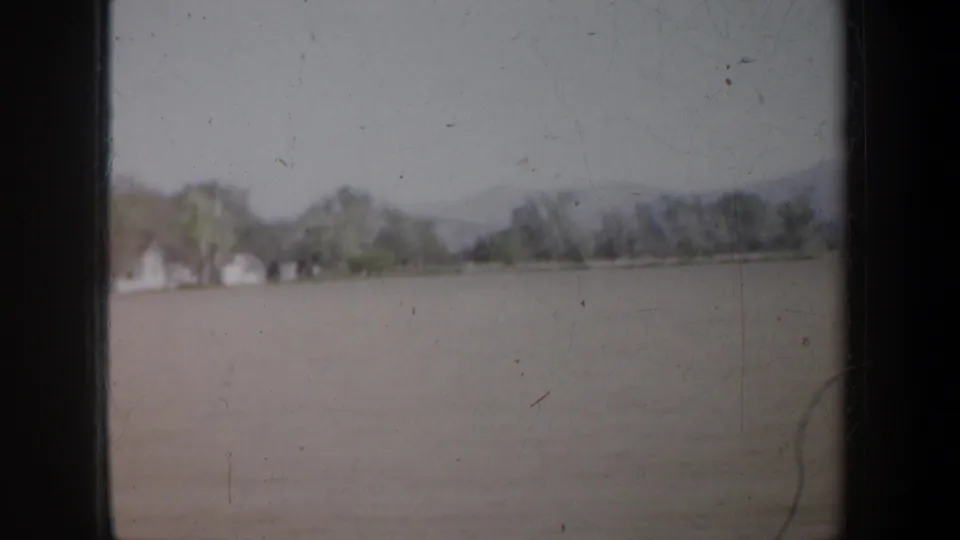 1957 Oregon home-movie frame showing open water or flat terrain with distant houses and tree line seen through the car window.