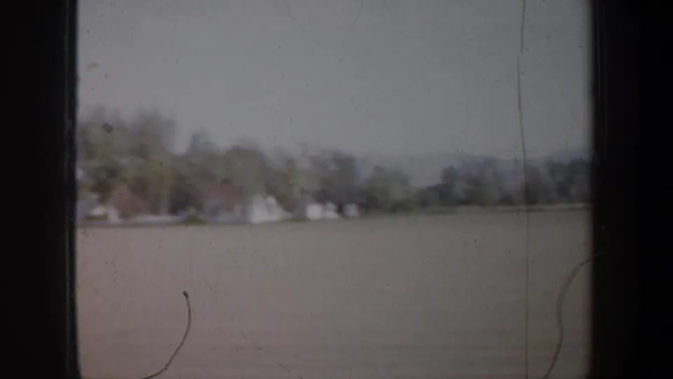 1957 Oregon frame showing distant houses, trees, and open water or flat ground from a moving car-window perspective.