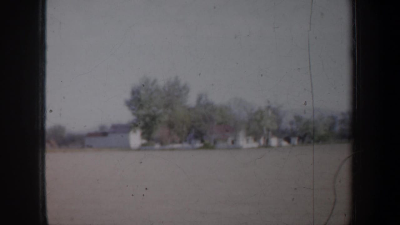 Soft roadside view of open land, distant pale buildings, trees, and a hazy sky through dark film borders.