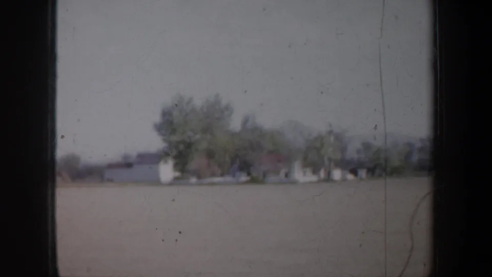 1957 Oregon 8mm frame showing open rural ground, pale farm buildings, green trees, and distant low hills from a moving vehicle.