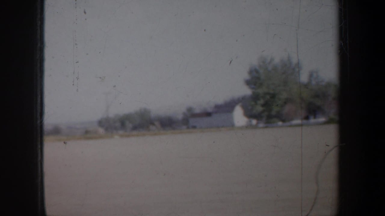 White barn-like building and green trees beside open land, seen through scratched 8mm film edges.