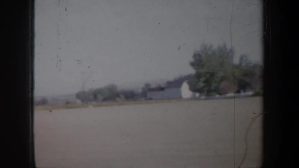 1957 Oregon roadside view of a white barn-like structure, trees, open ground, and hazy hills in a soft 8mm frame.