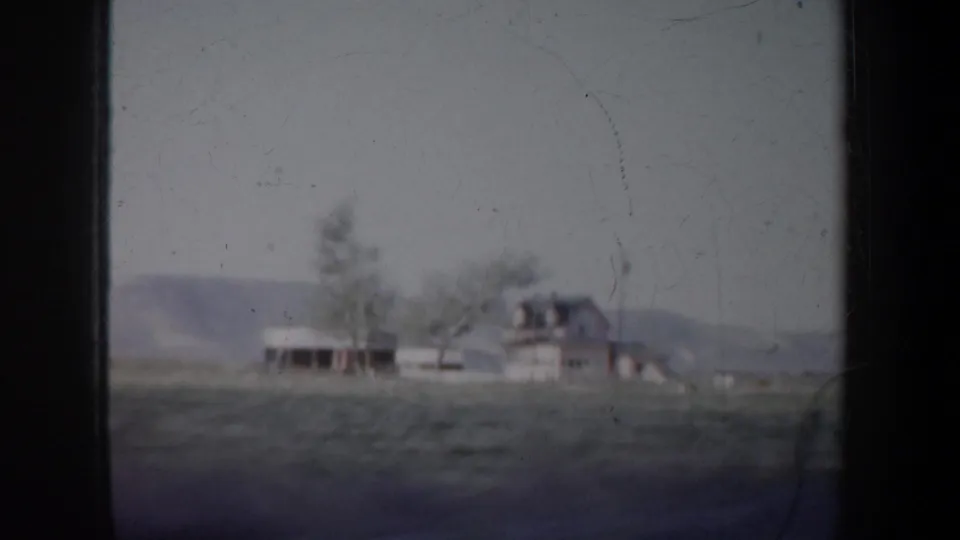 1957 Oregon frame showing a small rural building cluster, trees, open land, and low hills from a moving 8mm home movie camera.