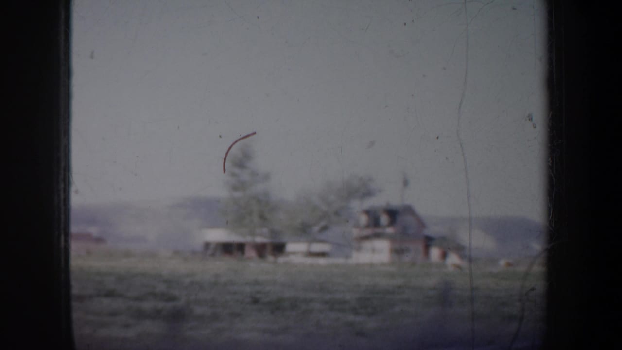 Distant farmhouse and trees across a field, framed by dark 8mm film borders.