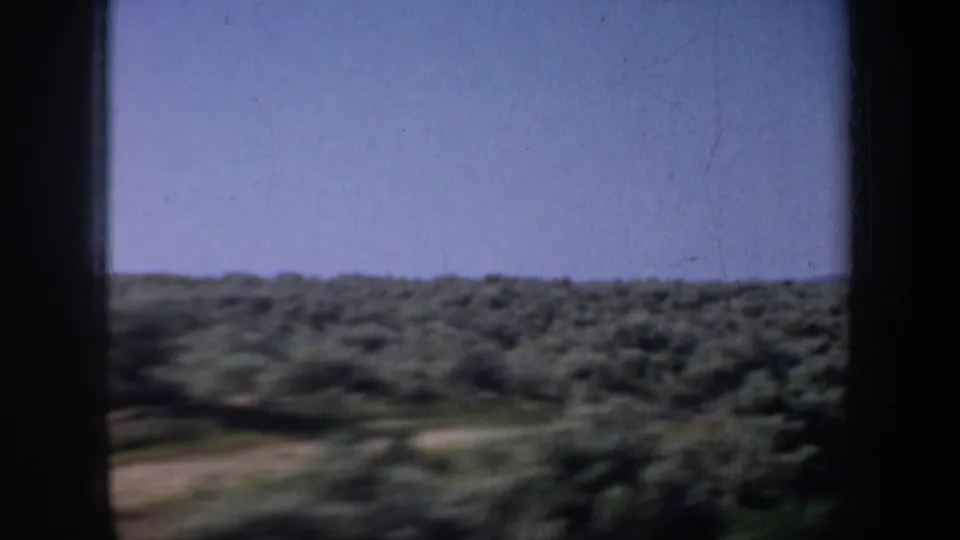 1957 Oregon 8mm landscape frame showing open scrubland, low hills, and sky from a moving road-view perspective.