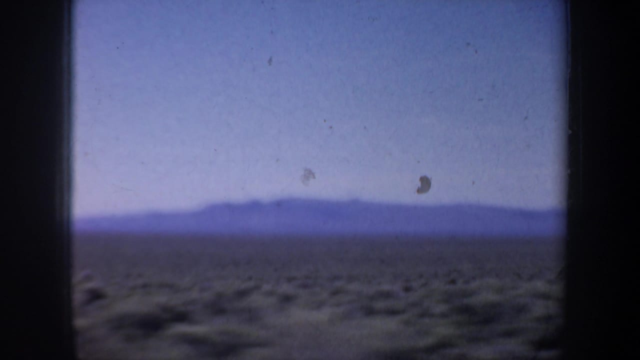 1957 Oregon 8mm frame with open brown fields and a distant mountain or hill silhouette under hazy blue sky.