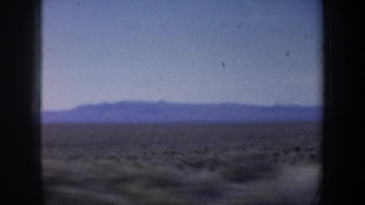 1957 Oregon 8mm view of low shrubs across an arid plain, with hazy hills on the horizon and dark film edges.