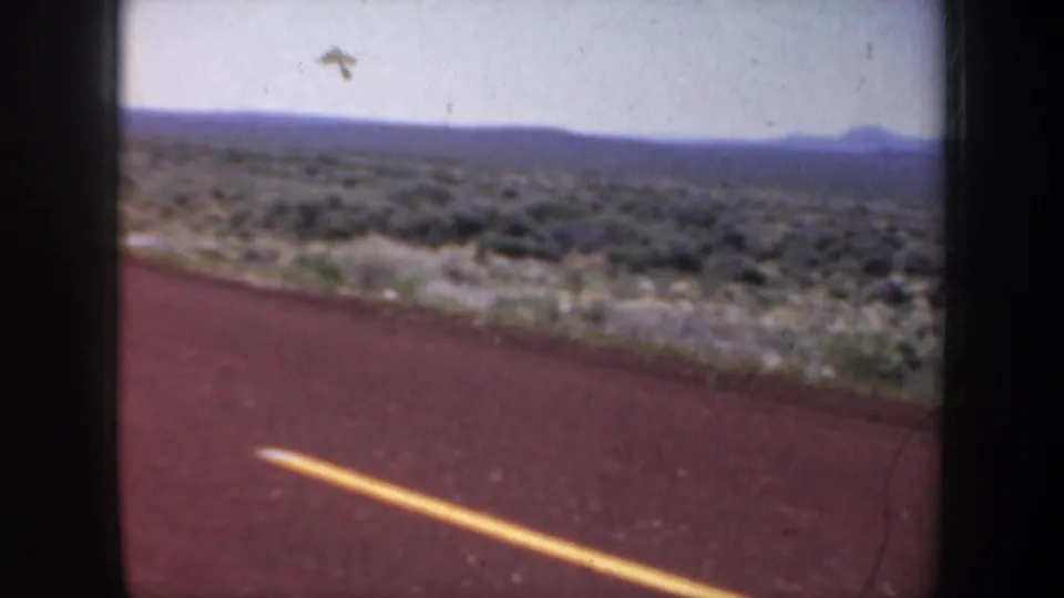1957 Oregon roadside view from a moving vehicle, with empty pavement, yellow line, scrubland, and distant hills.