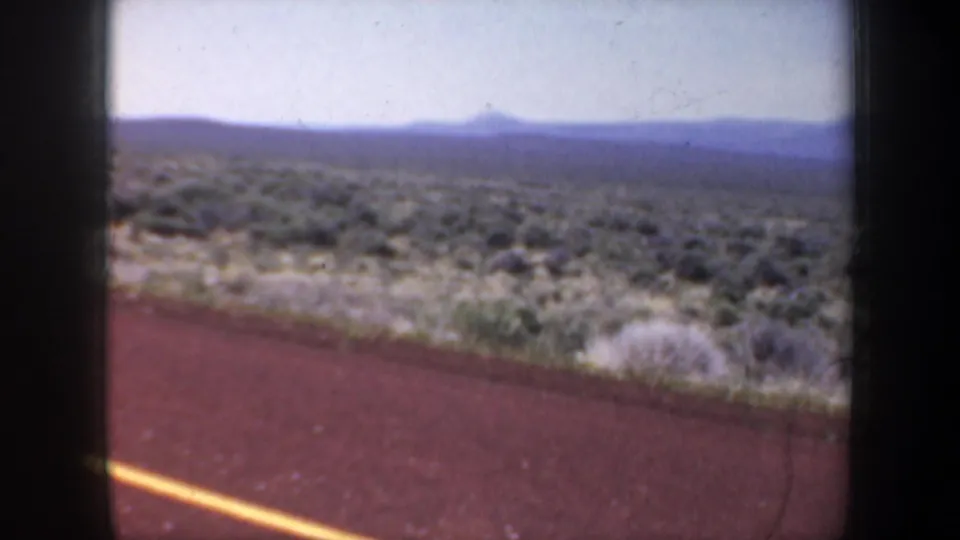 1957 Oregon 8mm travel frame with the road edge in the foreground and scrub-covered terrain stretching to distant hills.