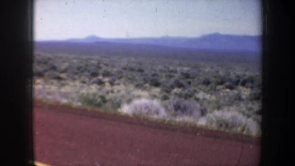 1957 Oregon 8mm frame showing open roadside scrub and distant mountains, filmed from a moving vehicle.