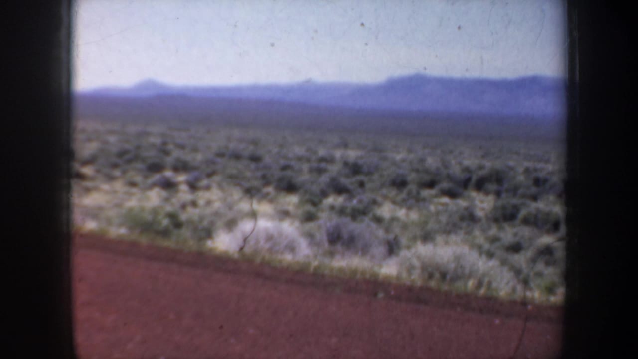 Arid scrubland with distant mountains under a pale sky, seen beside a road edge.