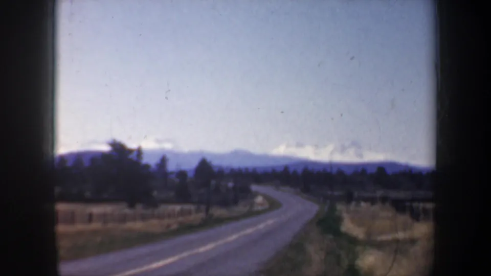 1957 Oregon 8mm frame showing a curving rural road, dry fields, fence lines, trees, and distant snowy mountains.