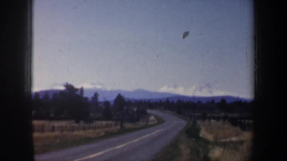 1957 Oregon road scene with a two-lane curve, roadside fields, dark tree line, and distant mountain horizon.