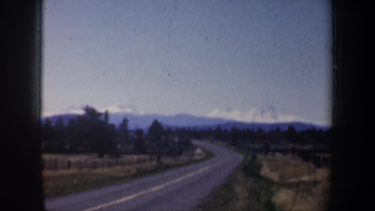 1957 Oregon 8mm view from a moving vehicle showing a curving road, dry fields, trees, and distant blue-gray mountains.