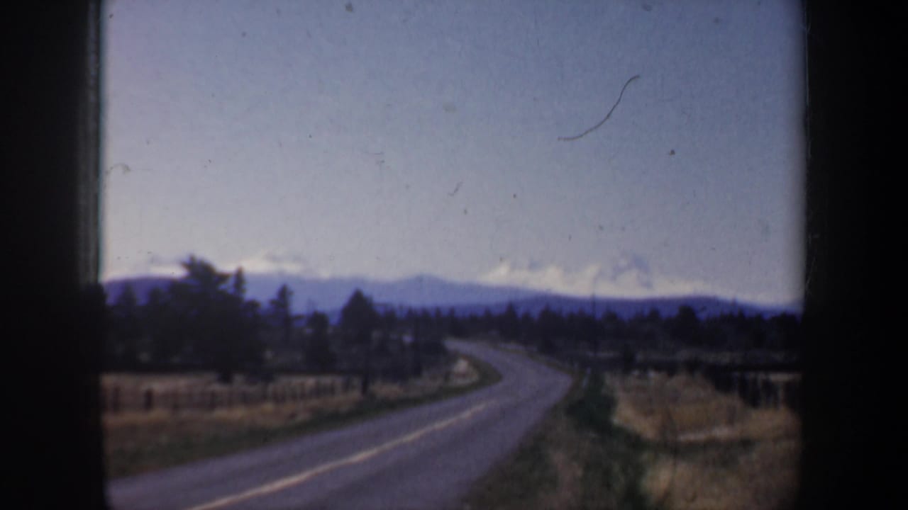 Oregon home-movie frame with a two-lane road curving through open fields toward trees and snow-lined distant mountains.