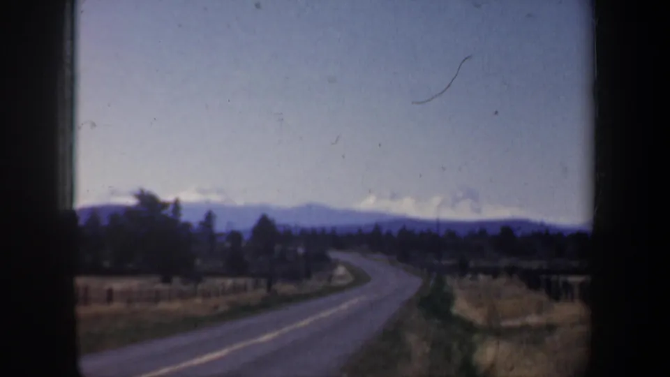 Oregon home-movie frame with a two-lane road curving through open fields toward trees and snow-lined distant mountains.