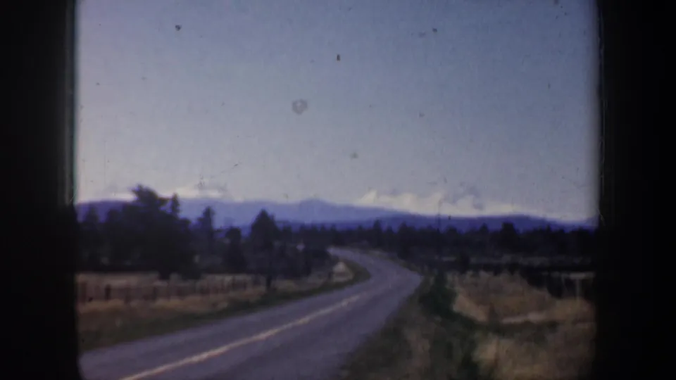 Soft 8mm frame of rural Oregon road travel, with tan fields, dark tree line, and blue-gray mountains below a pale sky.