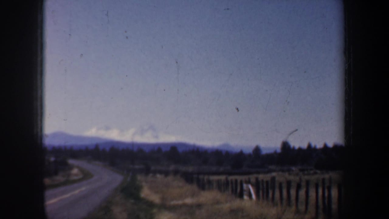 Winding rural road beside fence posts with distant mountains under a blue sky.
