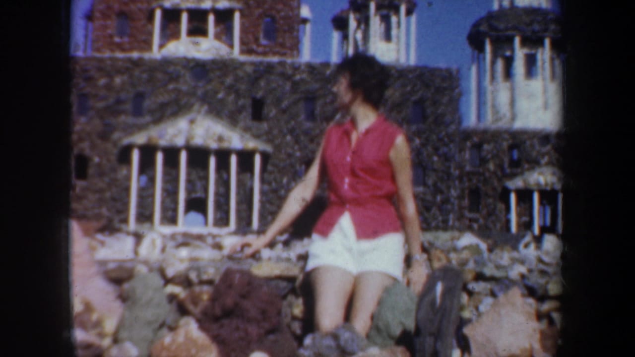 Woman seated on rocks in front of a stone building with columns and towers.