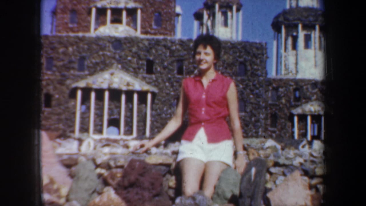 The woman smiles toward the camera in a 1957 Oregon travel scene, with rocks, a stone facade, and a small Statue of Liberty replica visible.