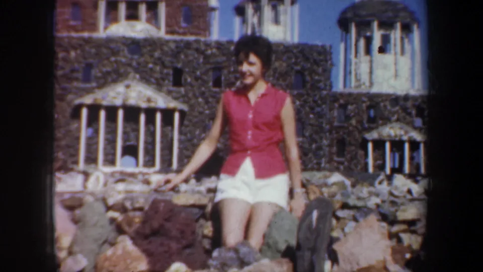 A woman in a red sleeveless top sits among rocks at an Oregon tourist setting, smiling in soft color 8mm footage from 1957.