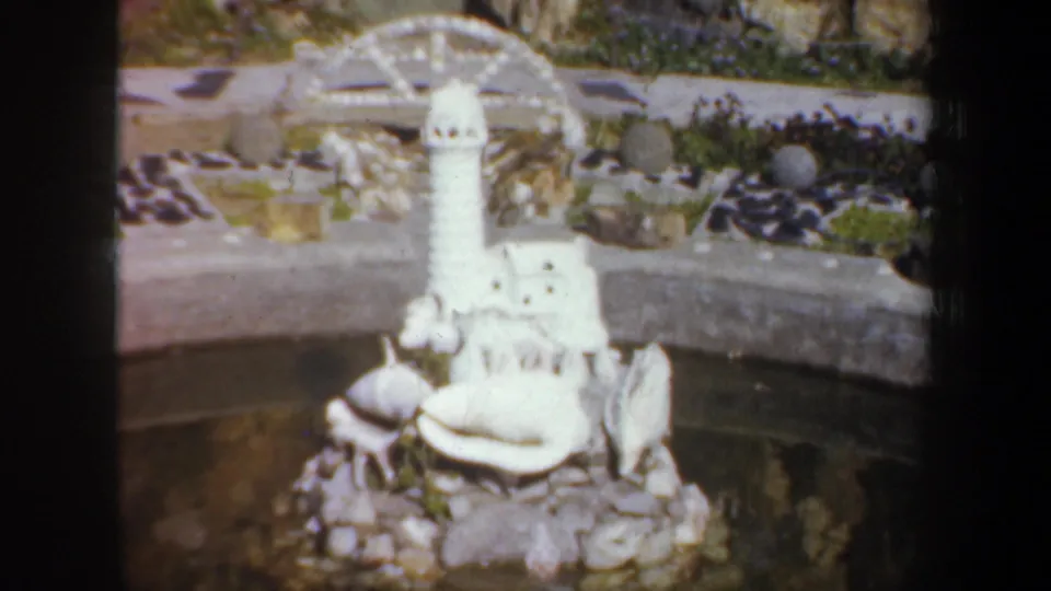 1957 Oregon 8mm frame showing a white miniature lighthouse set among shells, rocks, and pond water in a garden display.