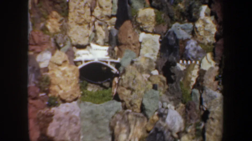 1957 Oregon 8mm frame showing a small arched bridge embedded in varied rocks within the miniature garden display.