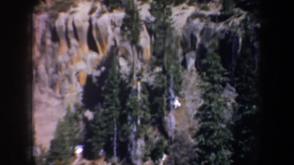 1957 Oregon scenic frame showing a rocky hillside, evergreen trees, dark woods, and small bright distant shapes that are not identifiable.