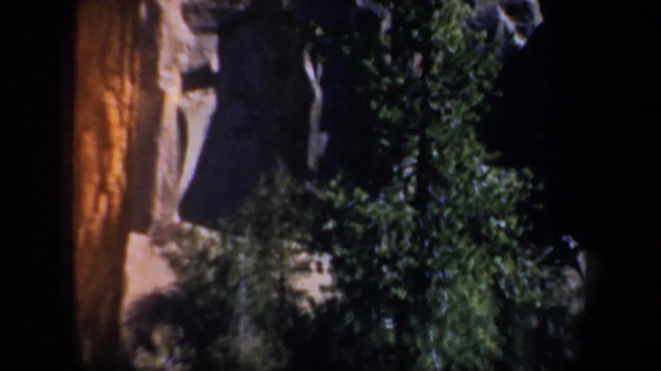 1957 Oregon 8mm frame with a shadowed rock wall, evergreen trees, and dense brush in a dark outdoor view.
