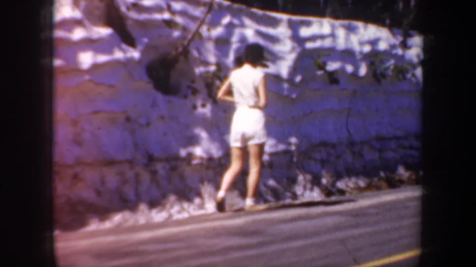 A lone woman in white walks away along an Oregon roadside beside a pale stone wall in this 1957 home-movie frame.