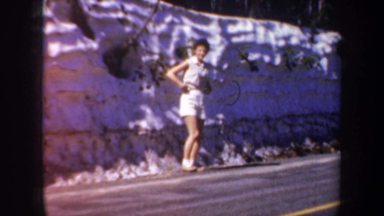 Woman in white sleeveless top and shorts poses beside a sunlit roadside wall.