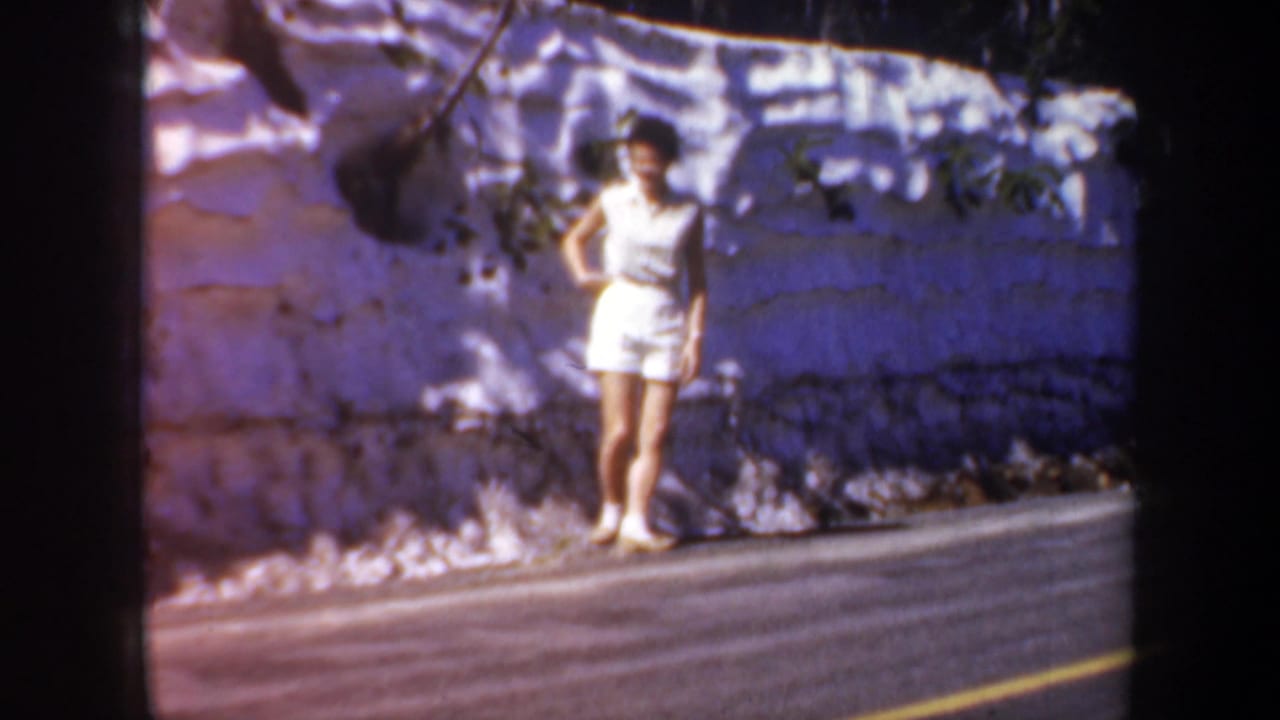 Woman stands near a roadside wall with bright sunlight and shadow patterns.