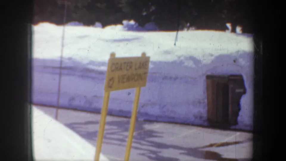 A yellow Crater Lake Viewpoint sign appears beside snowbanks, pavement, and a trash bin in the Oregon travel footage.