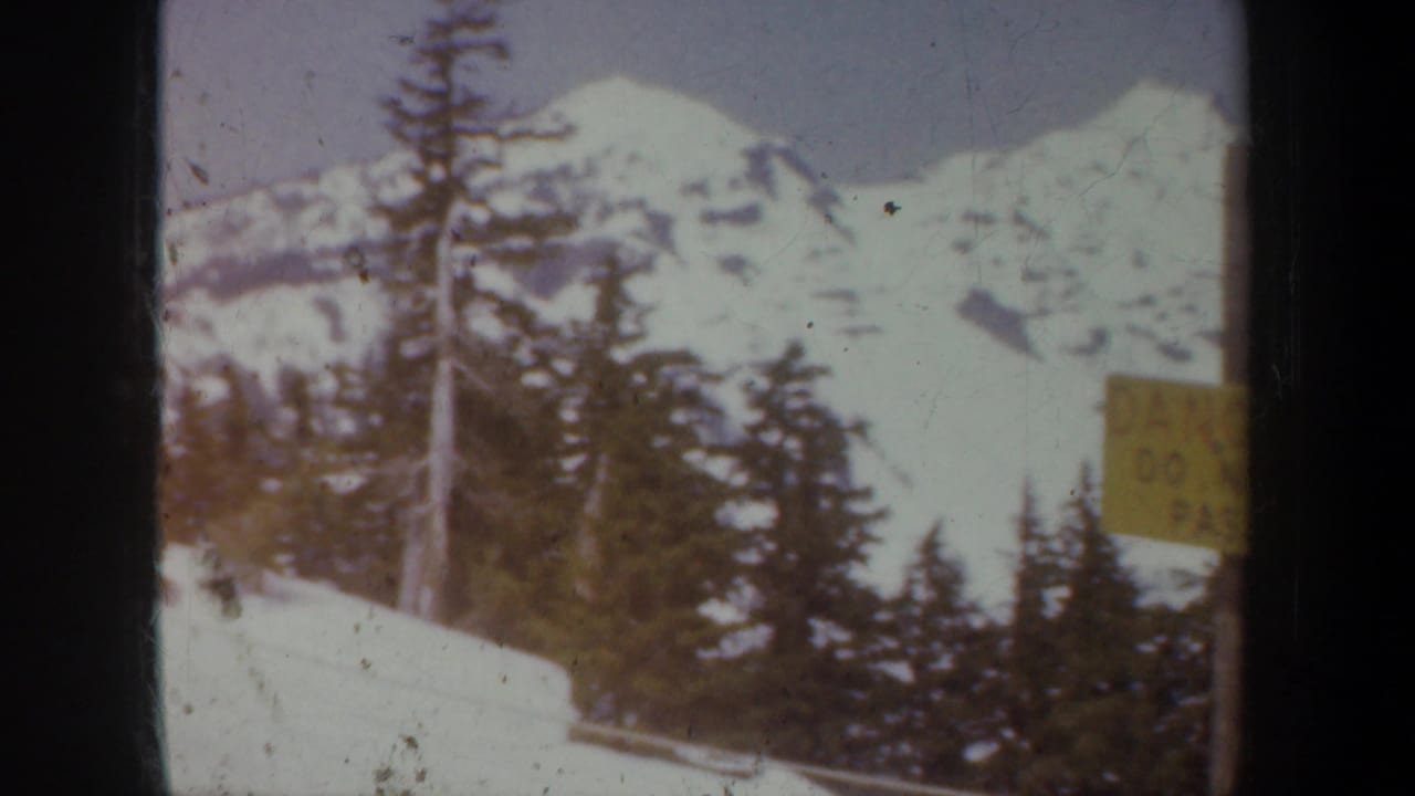 Snowy mountain slope with evergreen trees and a yellow danger sign near the edge of the frame.