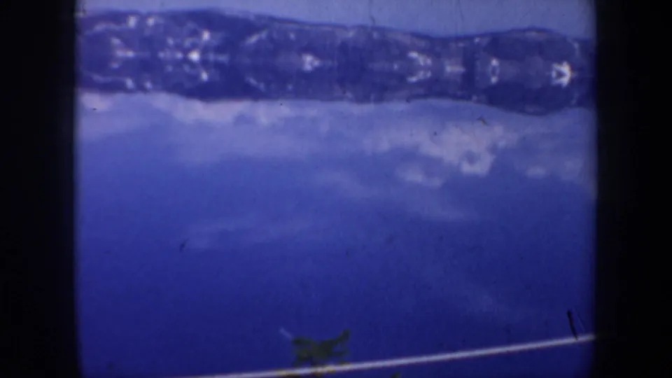 1957 Oregon 8mm frame showing a blue lake reflection, snowy ridge, and a small dark foreground shape near the lower edge.