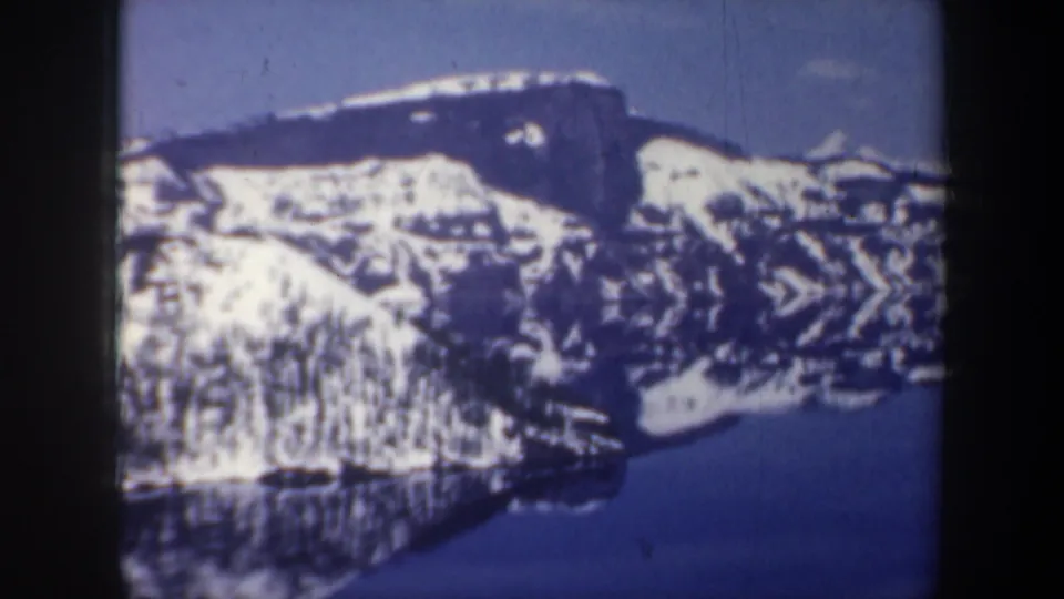1957 Oregon 8mm frame showing a broad snow-covered mountain reflected in still blue water.