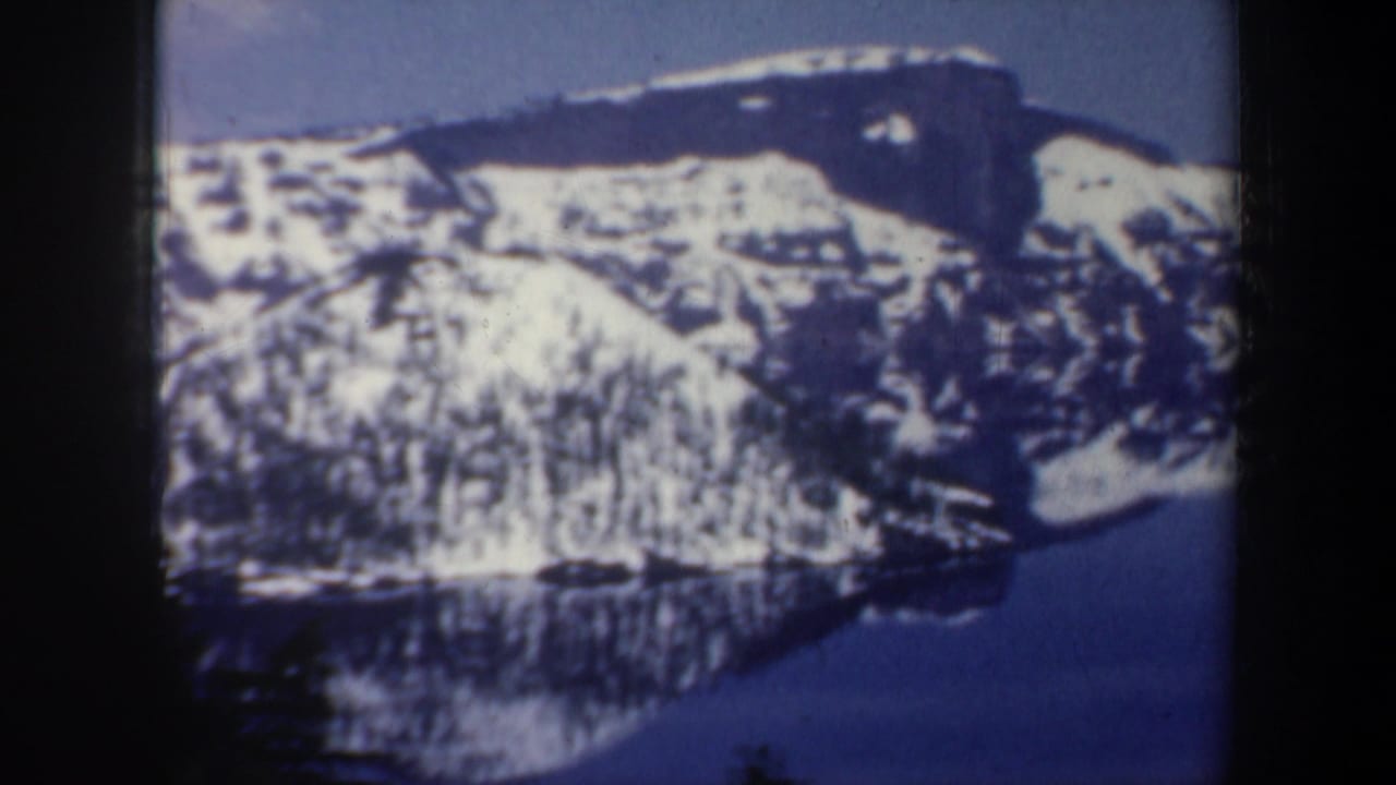 Snow-covered mountain ridge above a dark lake with trees in the foreground.